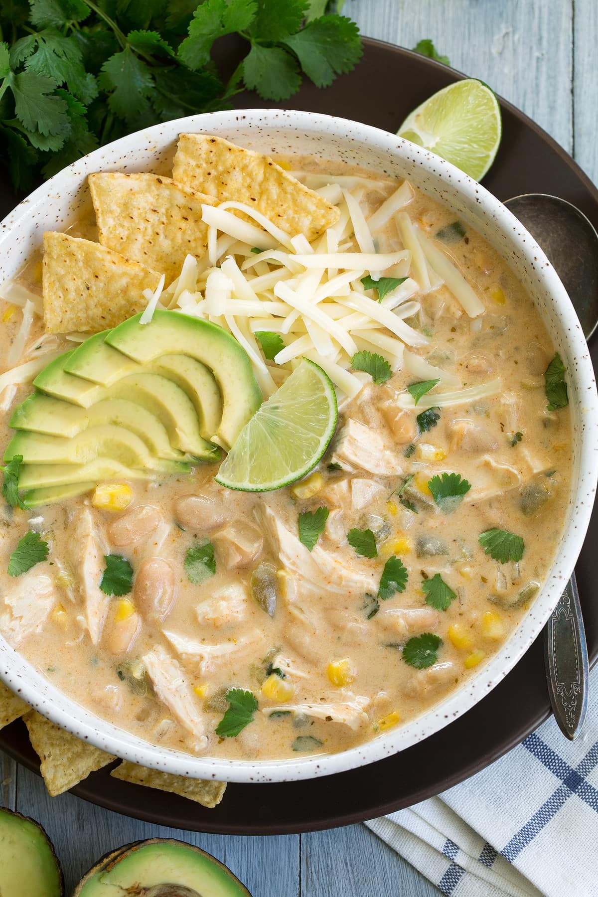 White Chicken Chili Overhead image of white chicken chili in a large white bowl sitting on a brown plate. Chili is garnished with cheese, chips, avocado, cilantro and lime.